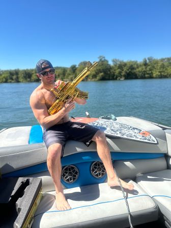Shirtless man wearing sunglasses and a backwards cap sitting on a boat at a sunny lake, holding a shiny gold inflatable toy rifle beside a wakeboard under a clear blue sky.