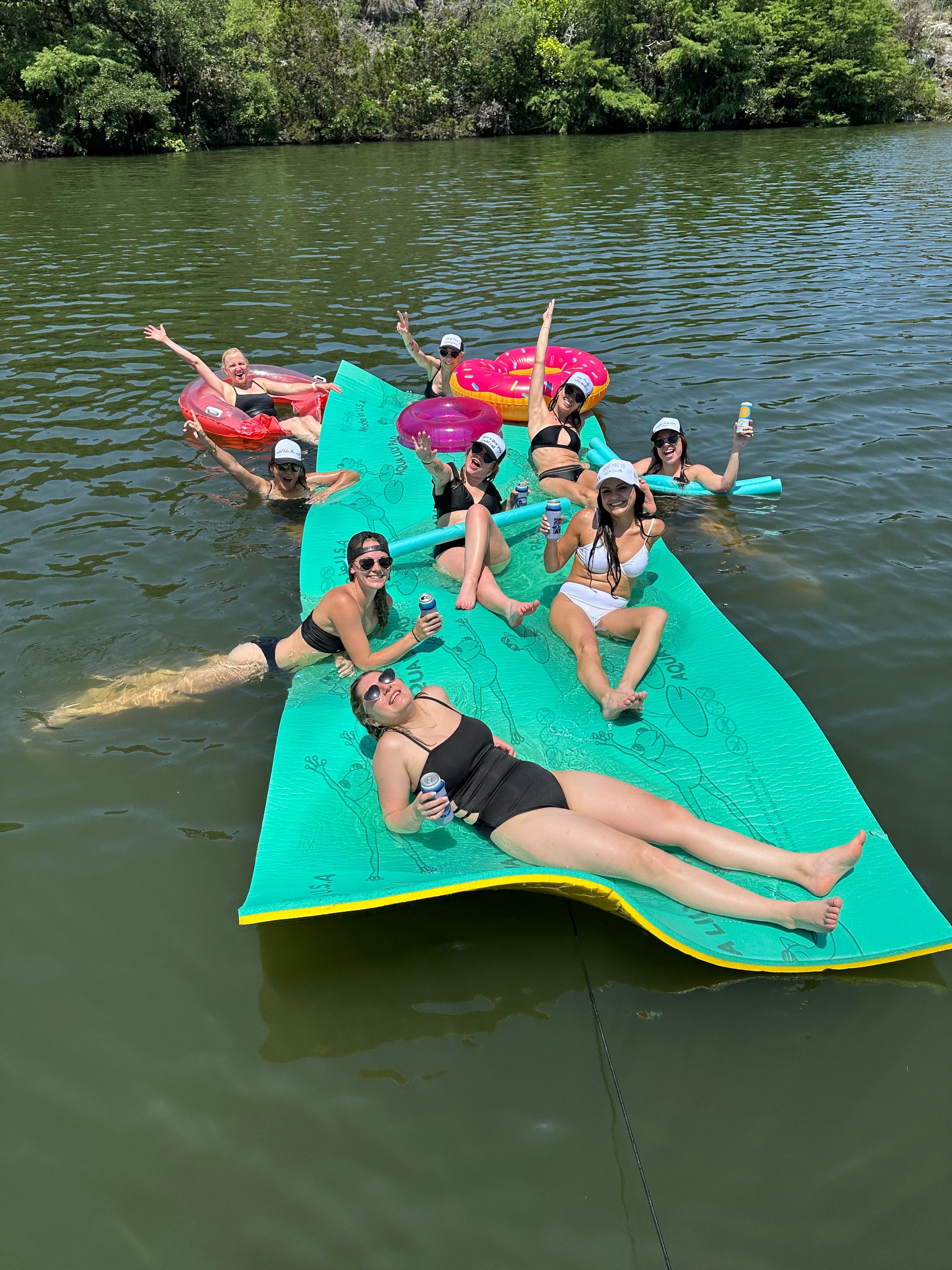 Group of friends lounging on a large green floating mat with colorful inflatables in a sunny lake, holding drinks near a tree-lined shore.