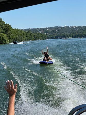 Two people in life jackets enjoying summer lake tubing on a blue inflatable towable, bouncing on a boat wake with tree-lined shoreline and rolling hills under a clear blue sky.