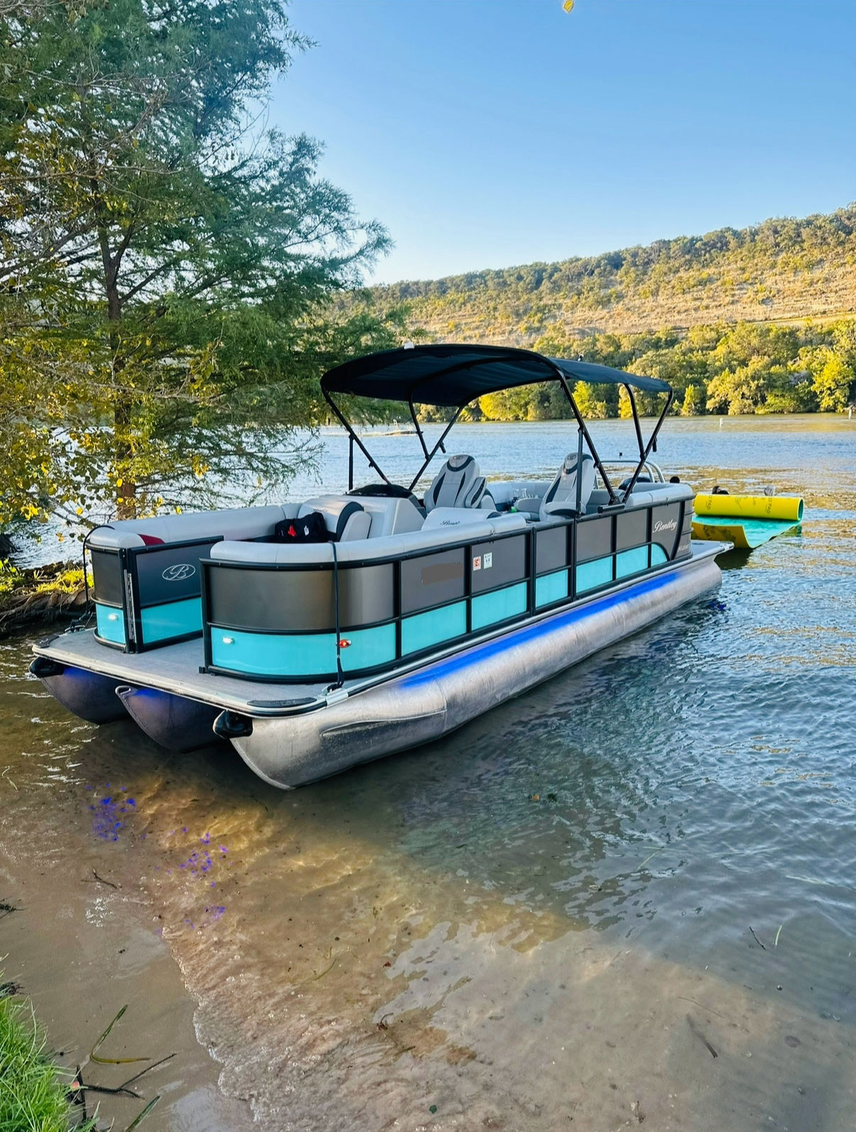 Turquoise-accented pontoon boat with black canopy beached on a sandy lake shore, calm water with a yellow inflatable, and tree-lined hillside in the background