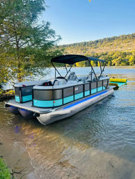Turquoise-accented pontoon boat with black canopy beached on a sandy lake shore, calm water with a yellow inflatable, and tree-lined hillside in the background