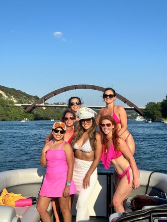 Six women in bright pink swimwear smiling and posing on a boat on Lake Austin, Texas, with the Pennybacker (360) Bridge arching over the water under a clear blue sky.