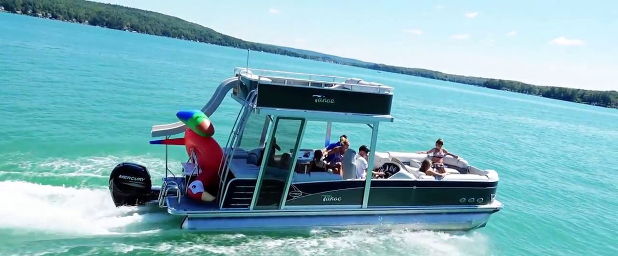 Sunny summer pontoon boat cruising on bright turquoise lake, group of people relaxing onboard and a giant inflatable parrot at the stern with tree-lined shore in the distance.