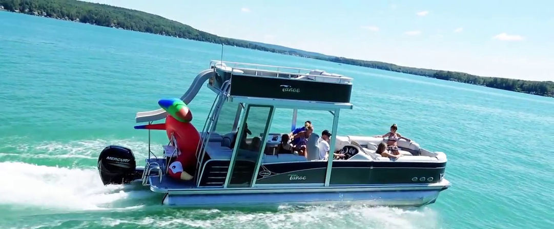 Sunny summer pontoon boat cruising on bright turquoise lake, group of people relaxing onboard and a giant inflatable parrot at the stern with tree-lined shore in the distance.