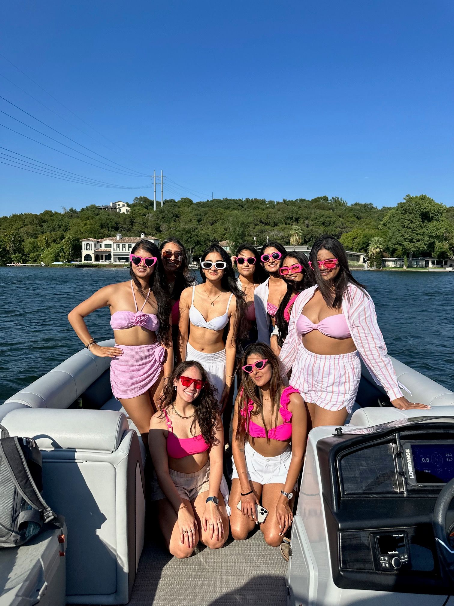 Group of women in pink and white swimwear and sunglasses posing on a pontoon boat on a sunny lake with tree-lined shoreline and clear blue sky — summer lake boat party.