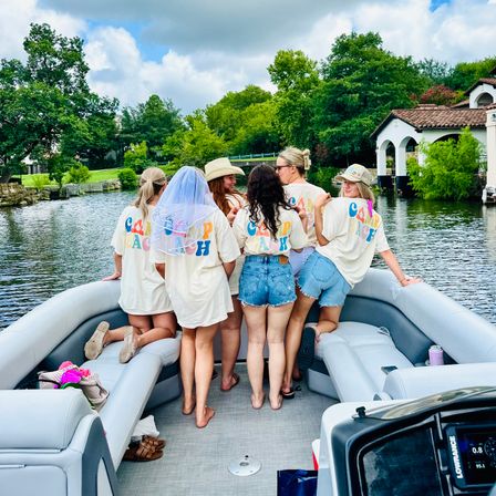 Friends in matching colorful-letter shirts and denim on a pontoon boat, celebrating a bachelorette with a veil while cruising a tree-lined lake past waterfront homes on a cloudy summer day
