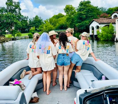 Friends in matching colorful-letter shirts and denim on a pontoon boat, celebrating a bachelorette with a veil while cruising a tree-lined lake past waterfront homes on a cloudy summer day
