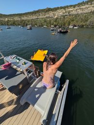 Summer lake scene: person in a bikini sitting on a boat slide with arm raised, overlooking a yellow inflatable mat with swimmers, multiple boats on the water and a rocky cliff shoreline under a clear blue sky.