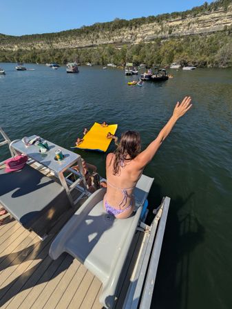 Summer lake scene: person in a bikini sitting on a boat slide with arm raised, overlooking a yellow inflatable mat with swimmers, multiple boats on the water and a rocky cliff shoreline under a clear blue sky.