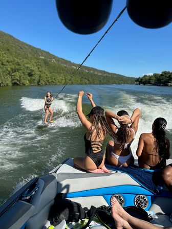 Three friends in swimsuits sitting on a motorboat watching a woman wakesurfing in the boat’s wake on a sunny lake with tree-lined hills