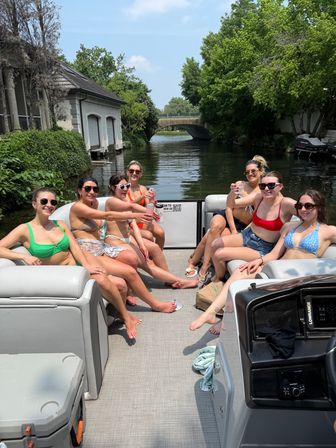 Eight friends in swimsuits toasting drinks on a pontoon boat cruising a tree-lined canal on a sunny summer day, with waterfront houses and a small bridge ahead.