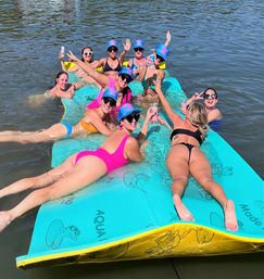 Group of women in colorful swimsuits and matching blue caps lounging and cheering with canned drinks on a large turquoise floating mat in a calm lake
