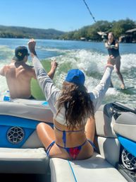 Friends on a boat cheering as a wakesurfer rides the wake on a sunny lake — woman in a blue cap and bikini raises her arms while a shirtless friend steers.