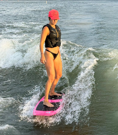 Person in a black life vest and pink cap wakesurfing on a bright pink board, balancing on a small wave in open lake water during golden-hour light.