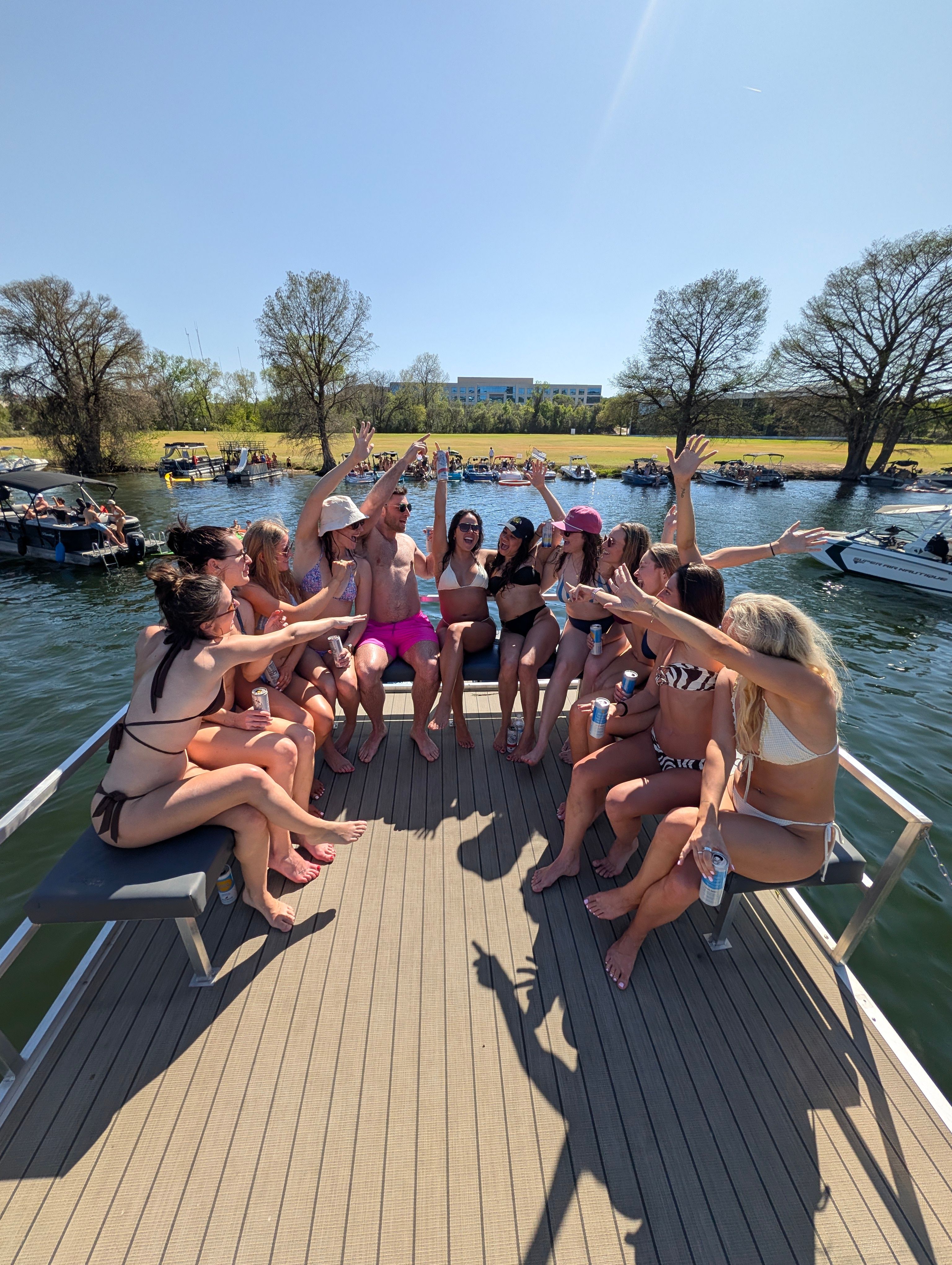 Group of people in swimsuits cheering with drinks on a pontoon boat during a sunny lakeside summer boat party, other boats anchored near a tree-lined shore under a clear blue sky.