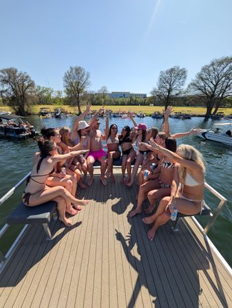 Group of people in swimsuits cheering with drinks on a pontoon boat during a sunny lakeside summer boat party, other boats anchored near a tree-lined shore under a clear blue sky.