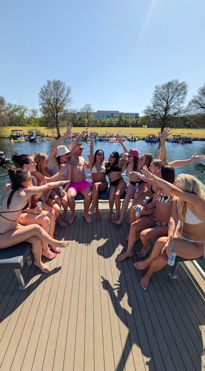 Group of people in swimsuits cheering with drinks on a pontoon boat during a sunny lakeside summer boat party, other boats anchored near a tree-lined shore under a clear blue sky.