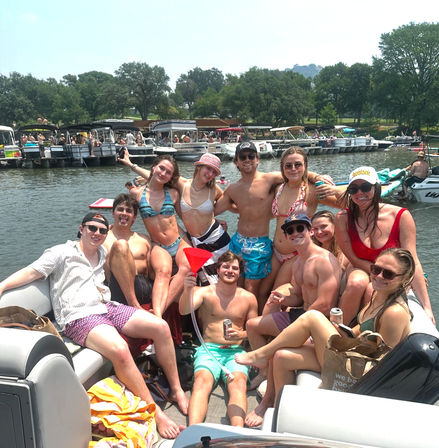 Group of young adults in swimsuits enjoying a sunny lake party on a pontoon boat, smiling and holding drinks with a busy dock and tree-lined shore in the background.