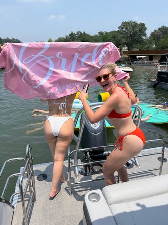 Two women on a pontoon boat at a sunny lake bachelorette celebration — one holds a pink 'Bride' towel over her white bikini, the other in a red bikini laughs and points at the camera, with floating mats and other boats in the background.
