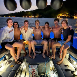 Seven friends sitting together on the back of a lit boat at dusk on a lake, wearing swimwear and casual summer clothes, with shoreline lights and lakeside houses glowing in the background and a cooler with drinks in the foreground.