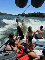 Friends on a motorboat cheering as a wakesurfer rides the boat’s wake on a sunny lake with tree-lined shoreline — summer water sports scene.