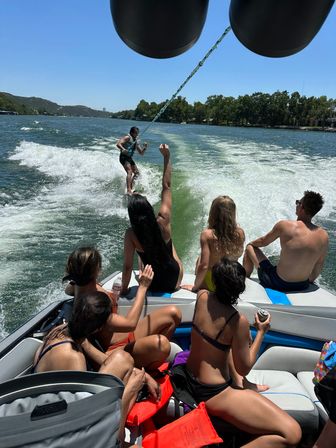 Friends on a motorboat cheering as a wakesurfer rides the boat’s wake on a sunny lake with tree-lined shoreline — summer water sports scene.