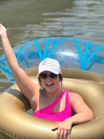 Smiling person in a pink one-shoulder swimsuit and white cap, sunglasses raised arm while lounging in a gold inflatable tube and clear blue-ring float on a calm lake — summer lake fun.