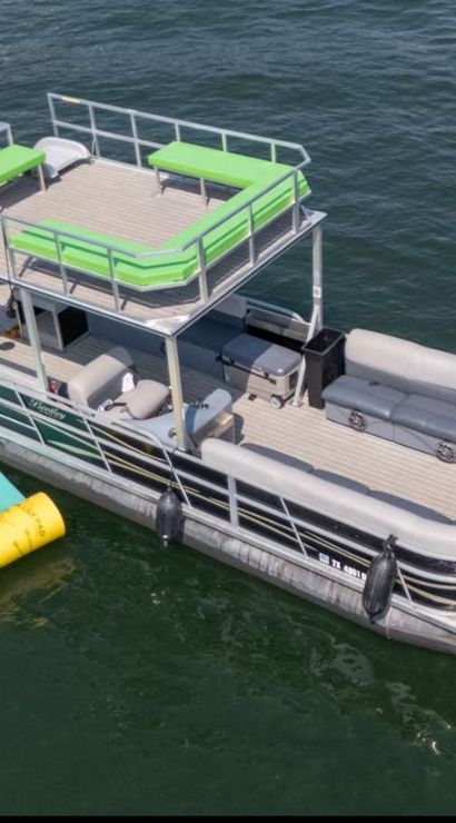 Aerial view of a double-decker pontoon boat with lime-green cushions and upper sun deck on a calm lake, docked beside bright yellow and teal inflatable water mats.
