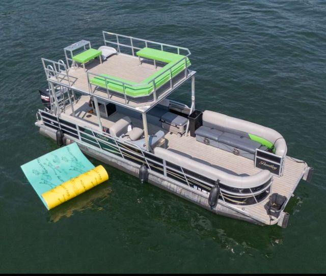 Aerial view of a double-decker pontoon boat with lime-green cushions and upper sun deck on a calm lake, docked beside bright yellow and teal inflatable water mats.