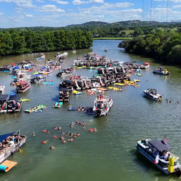 Aerial view of a lively summer boating party on a sunlit river — clustered pontoon and motorboats with swimmers and colorful rafts floating nearby.