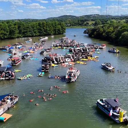 Aerial view of a lively summer boating party on a sunlit river — clustered pontoon and motorboats with swimmers and colorful rafts floating nearby.