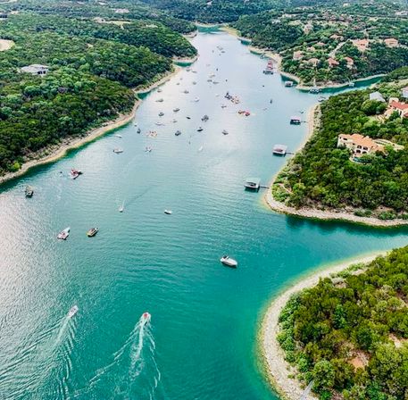 Aerial view of a turquoise lake inlet with boats and jet skis dotting the water, tree-covered shorelines, docks and scattered lakefront homes on a sunny day.