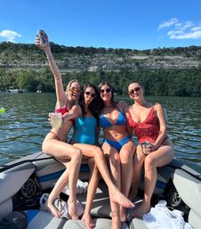 Four friends in colorful swimsuits sitting on a boat, raising drinks and smiling on a sunny lake with tree‑covered cliffs and other boats in the background.