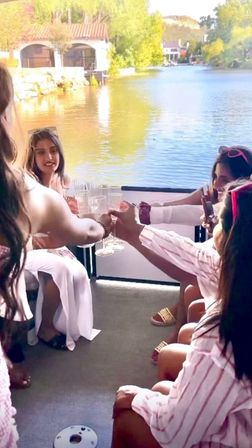 Sunlit lakeside toast on a boat — a group of women clinking champagne flutes with a calm lake and boathouses in the background, casual pink-and-white outfits and sunglasses.