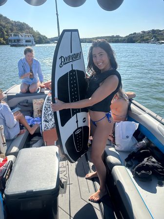 Smiling woman in a life vest holding a wakesurf board on a sunny summer lake boat, friends relaxing onboard and wooded shoreline in the background.