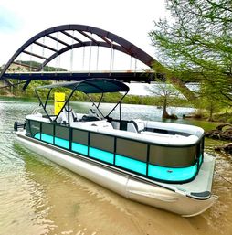 Teal-accent pontoon boat beached on a sandy riverbank beneath a large rust-colored arched bridge, framed by green shoreline trees and exposed roots, ready for a day on the water.