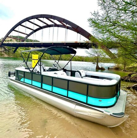 Teal-accent pontoon boat beached on a sandy riverbank beneath a large rust-colored arched bridge, framed by green shoreline trees and exposed roots, ready for a day on the water.