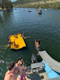Summer lake party with people lounging on a bright yellow floating mat and one person jumping from a dock slide, boats and wooded hills in the background.