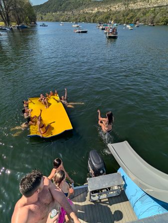 Summer lake party with people lounging on a bright yellow floating mat and one person jumping from a dock slide, boats and wooded hills in the background.