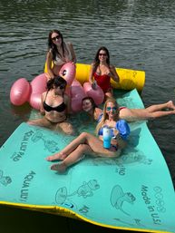 Five women in swimsuits relaxing on a turquoise floating mat and pink flamingo inflatable on a calm lake, holding drinks and enjoying a sunny summer day
