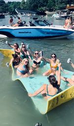 Friends cheering and raising cans while lounging on a large foam float in a crowded lake with anchored boats and tree-lined shore on a sunny summer day