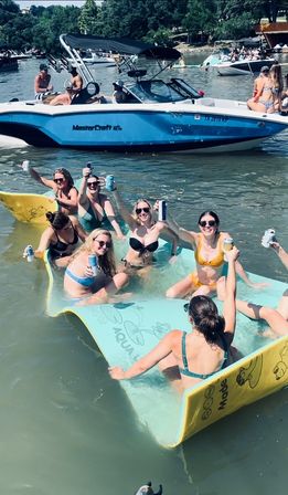 Friends cheering and raising cans while lounging on a large foam float in a crowded lake with anchored boats and tree-lined shore on a sunny summer day