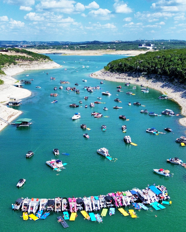 Aerial view of a busy summer lake cove with dozens of boats and a curved flotilla of colorful floating mats, swimmers in turquoise water between rocky green cliffs under a sunny blue sky.