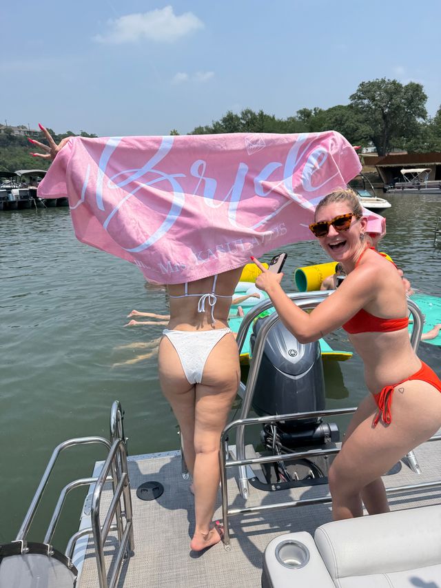 Two women laughing on a sunny pontoon boat at a lake — one in a white bikini holds a pink towel reading 'Bride' over her head while a friend in a red bikini points and smiles