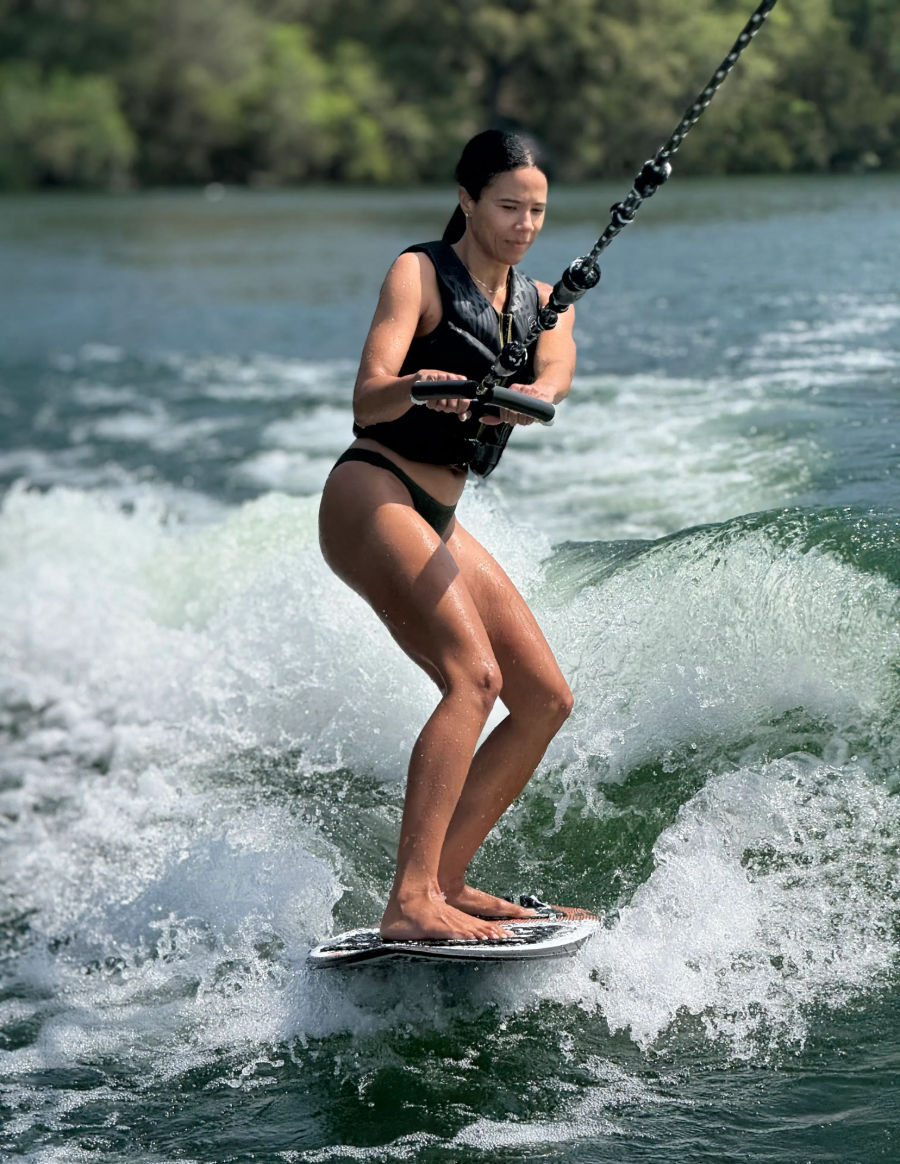 Person wakesurfing on a lake, barefoot on a small board holding a tow handle, wearing a black life vest and carving a foamy wake with tree-lined shore in the background.