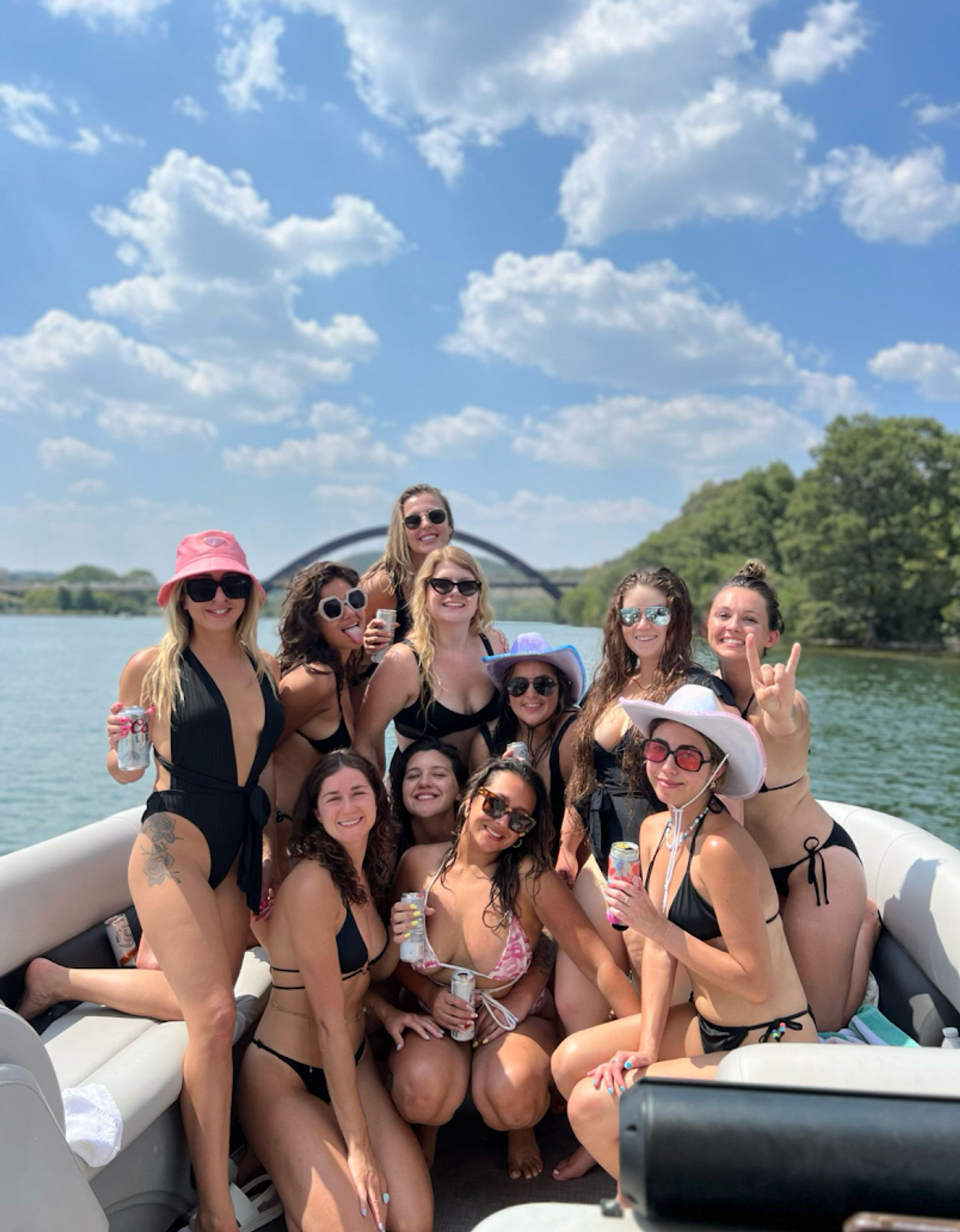Group of friends in bikinis smiling and holding drinks on a pontoon boat on a sunny summer lake, with a distant arched bridge and tree-lined shore under a blue sky with fluffy clouds.