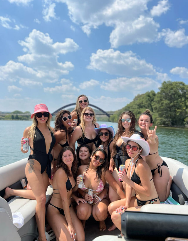 Group of friends in bikinis smiling and holding drinks on a pontoon boat on a sunny summer lake, with a distant arched bridge and tree-lined shore under a blue sky with fluffy clouds.
