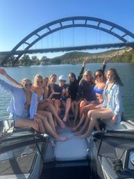 Cheerful group of friends posing and smiling on a boat at a sunny lake beneath a large arched bridge with wooded hills in the background.