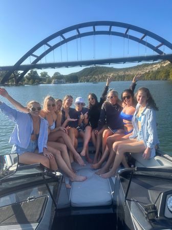 Cheerful group of friends posing and smiling on a boat at a sunny lake beneath a large arched bridge with wooded hills in the background.
