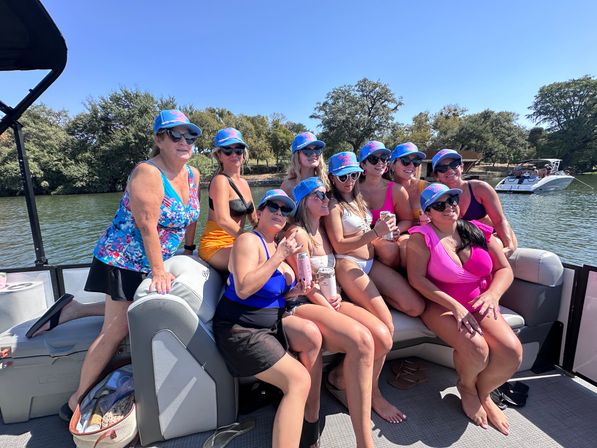 Cheerful group of women in colorful swimsuits and matching blue caps enjoying drinks on a pontoon boat on a sunny lake, with tree-lined shore and another boat in the background.